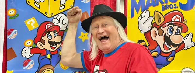 Charles Martinet sorrindo e com o punho erguido, entre painéis com o Mario