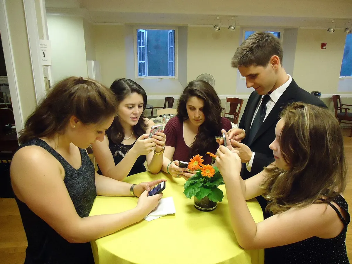 Cinco jovens em volta de uma mesa alta com toalha amarela, cada um olhando e tocando no próprio celular