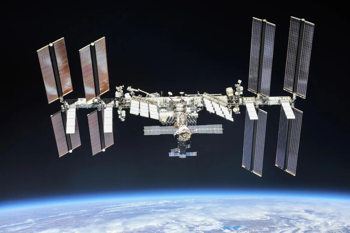 A Estação Espacial Internacional vista de fora, com os módulos no centro, os grandes painéis solares nas pontas e a curva da Terra embaixo