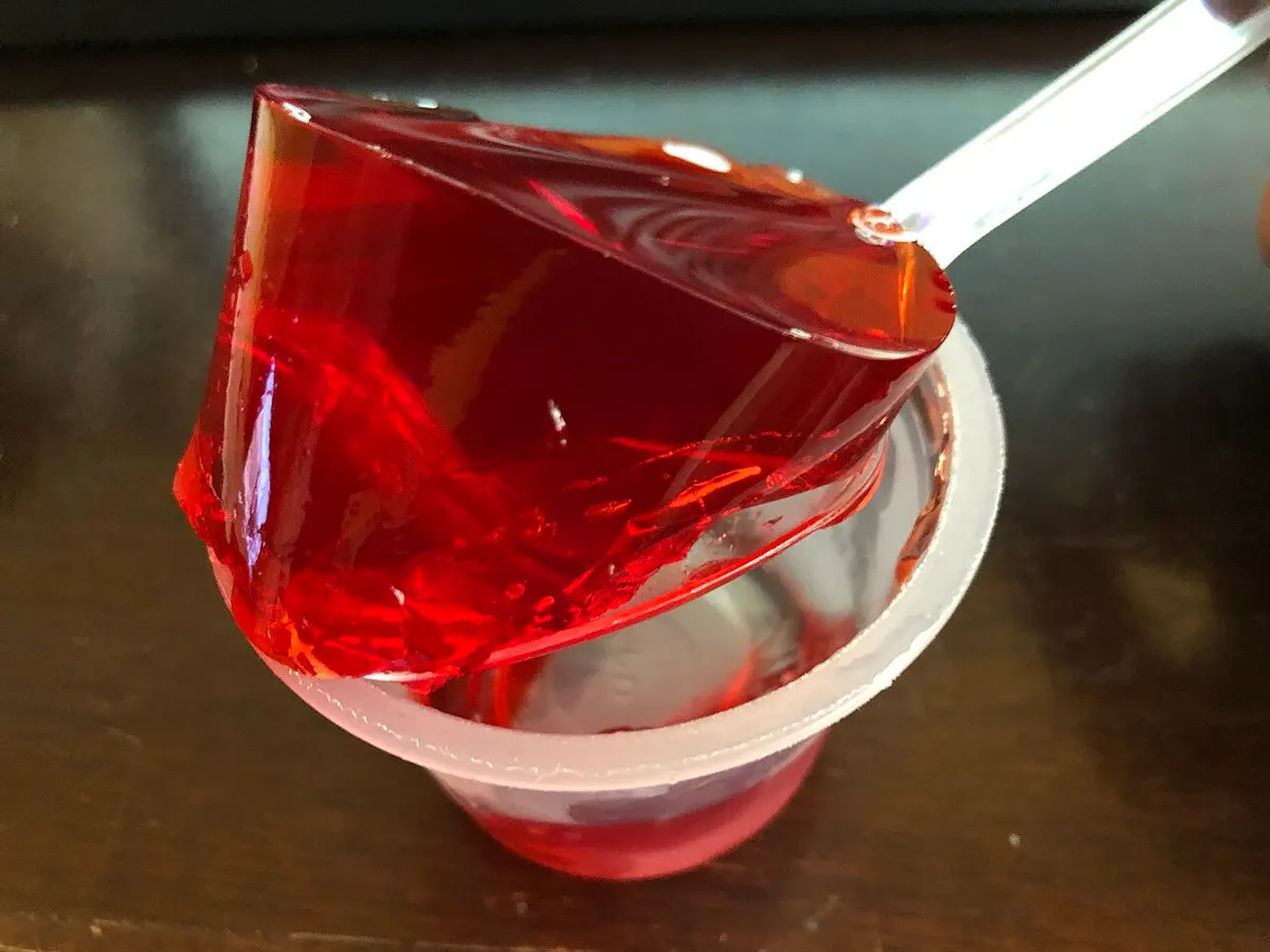 Gelatina vermelha de morango sendo levantada de um copinho plástico por uma colher, curvando-se sem se partir