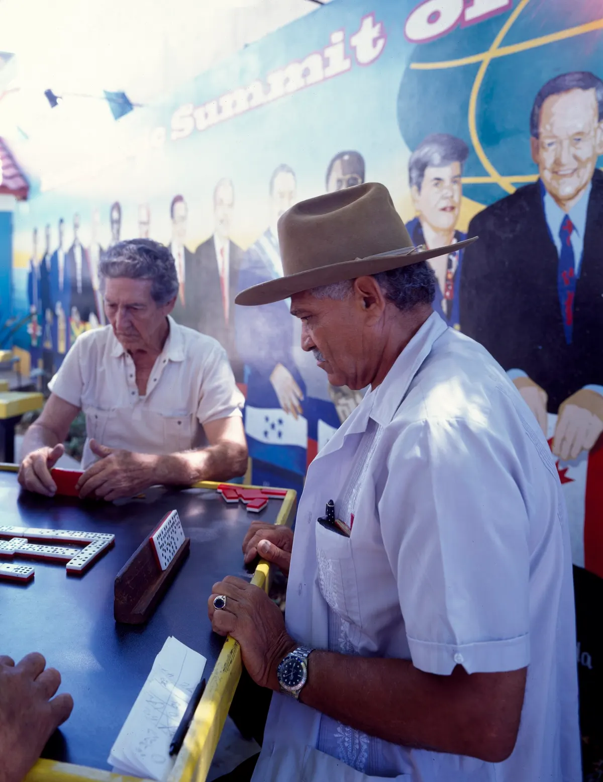 Dois homens idosos jogam dominó numa mesa ao ar livre, com pedras brancas na mesa e um suporte de madeira segurando as pedras de um jogador; ao fundo, um mural colorido