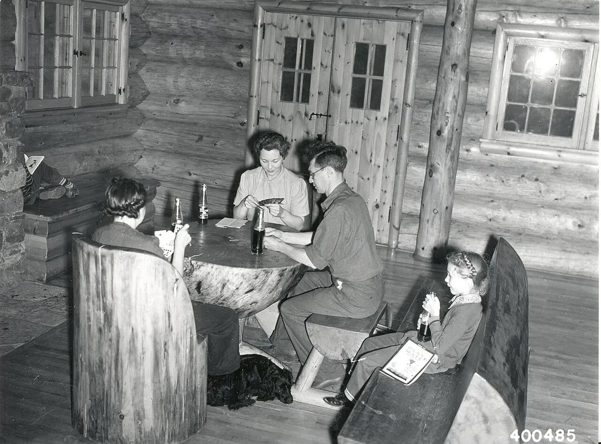 Foto em preto e branco de 1940: família joga cartas numa mesa de tronco dentro de uma cabana de madeira, com uma menina sentada no banco ao lado